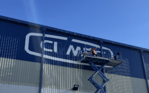 Hand-painted signage being applied to a corrugated metal warehouse, showcasing traditional branding techniques.