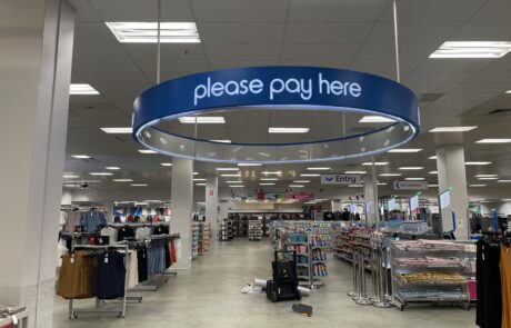 Circular 'Please Pay Here' suspended sign at a Kmart checkout area.